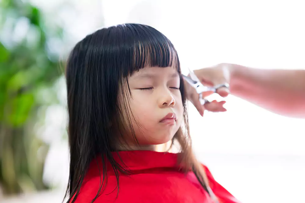 Kleines, asiatisches Mädchen mit geschlossenen Augen beim Friseur, während ihr Pony nachgeschnitten wird